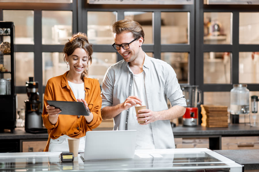 Two people reviewing a tablet beside a laptop in a cafe